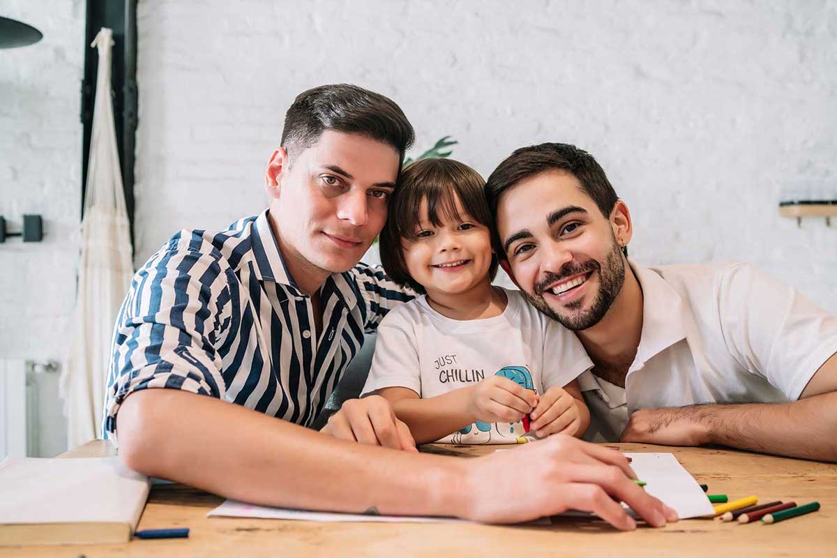 two young men with their young smiling adopted child all drawing together on the kitchen table