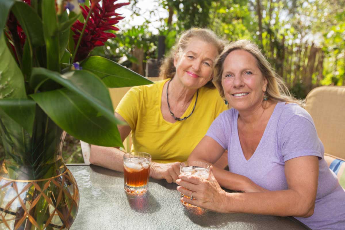 two mature women relaxing together at a table with tropic flowers and iced drinks